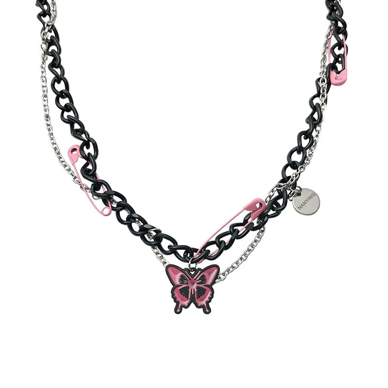 A gothic-inspired layered necklace featuring black and silver chains, pink enamel safety pins, and a pink butterfly pendant – edgy Y2K punk choker style.